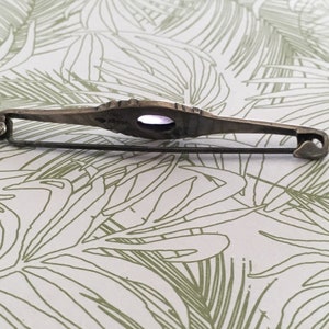 Op de afbeelding: Een zilveren broche met een paarse steen in een decoratief frame. De broche heeft een pin en een sluiting. De achterkant van de broche is gemarkeerd met "900".