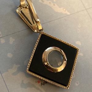 May include: A gold-toned pendant with a black square center and a circular, clear center. The pendant has a decorative bail and a diamond-shaped frame. The pendant is likely vintage or antique.