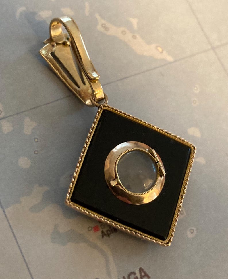 May include: A gold-toned pendant with a black square center and a circular, clear center. The pendant has a decorative bail and a diamond-shaped frame. The pendant is likely vintage or antique.