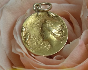 May include: A circular gold pendant featuring a relief design of a profile of a person with flowing hair and small birds. The pendant has a small jump ring attached to the top, allowing it to be worn on a chain or cord as a piece of jewelry.