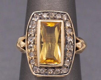 Puede incluir: Un anillo de oro con una gran gema amarilla rectangular en el centro, rodeada de gemas claras más pequeñas. El anillo tiene un diseño detallado y ornamentado con una estética vintage.