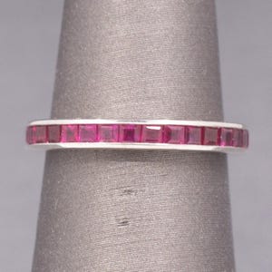 May include: A silver ring featuring a band of square-cut, bright pink gemstones. The gemstones are set closely together, forming a continuous line of colour. The ring is shown against a grey background.