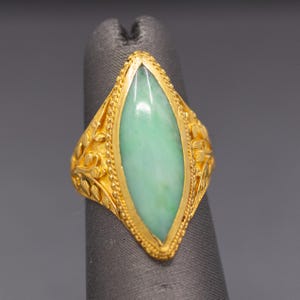 May include: A gold ring featuring a light green jade stone in a marquise shape. The ring has intricate floral and leaf detailing on the band. The jade stone is set in a gold bezel.