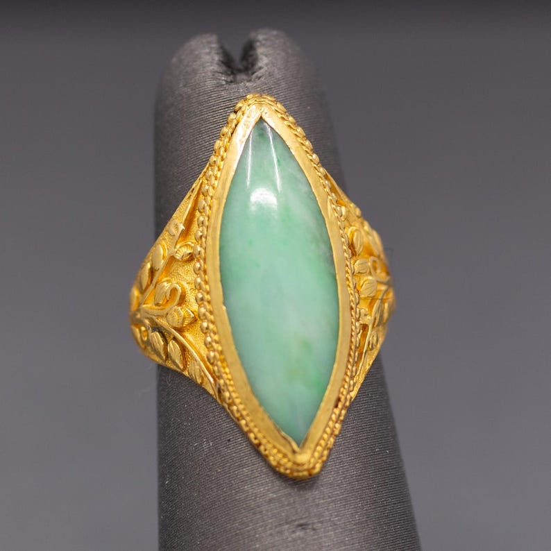 May include: A gold ring featuring a light green jade stone in a marquise shape. The ring has intricate floral and leaf detailing on the band. The jade stone is set in a gold bezel.