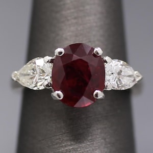 May include: A silver ring featuring a large oval-cut ruby center stone flanked by two pear-shaped diamonds. The ruby is a deep red color, and the diamonds are clear and sparkling. The ring is set on a dark gray background.