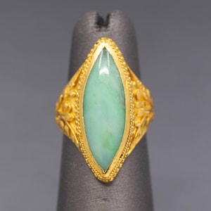 May include: A gold ring with a light green, oval-shaped stone. The ring has intricate floral designs on the sides and a textured border around the stone. The ring is displayed on a gray finger form.