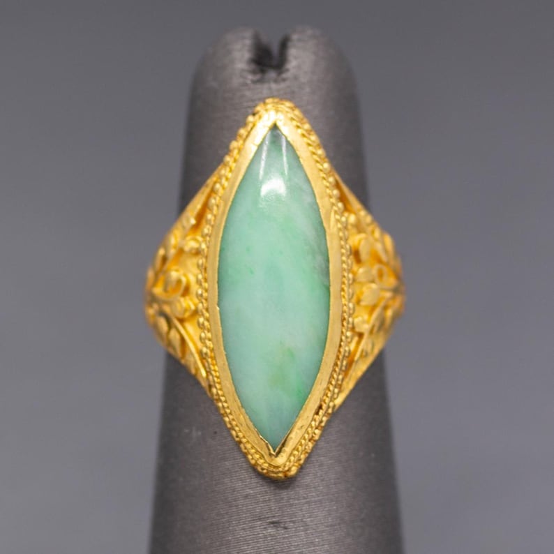 May include: A gold ring with a light green, oval-shaped stone. The ring has intricate floral designs on the sides and a textured border around the stone. The ring is displayed on a gray finger form.