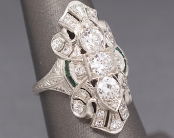 May include: An ornate, Art Deco-style ring featuring a central cluster of three large, sparkling diamonds. The ring is crafted in silver-toned metal and accented with smaller diamonds and emerald-green stones. The band has an engraved floral pattern.