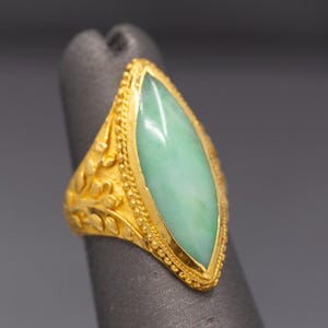May include: A gold ring featuring a marquise-cut, pale green jade stone. The ring has intricate filigree and beaded detailing around the stone and on the band. The ring is displayed on a gray surface.
