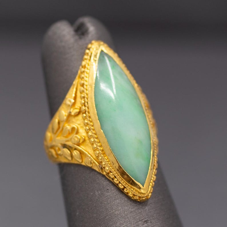 May include: A gold ring featuring a marquise-cut, pale green jade stone. The ring has intricate filigree and beaded detailing around the stone and on the band. The ring is displayed on a gray surface.