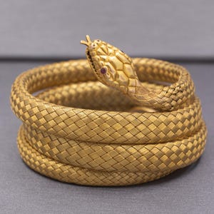 Pode incluir: Uma pulseira de serpente enrolada feita de metal dourado com uma textura trançada semelhante a uma malha. A cabeça da serpente apresenta gravações detalhadas de escamas e pequenos olhos de pedra vermelha incrustados, com uma língua bífida.