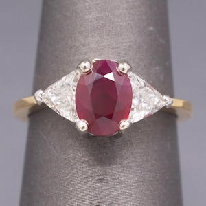May include: A gold ring featuring a large oval ruby center stone, flanked by two triangular diamonds. The ruby is a deep red color, and the diamonds are clear. The ring is set on a gold band.