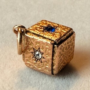 May include: A gold cube-shaped charm featuring a textured surface with an intricate pattern. The top face is set with a single blue gemstone, while the side face displays a clear, faceted stone centered within a star-shaped metal setting.