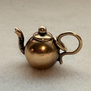 May include: A miniature gold-toned teapot charm featuring a rounded body, a curved handle, a spout with the text 14K engraved on it, and a small lid with a spherical knob. The charm includes a circular jump ring attached to the handle.