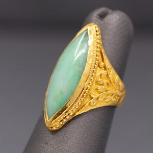 May include: A gold ring featuring a large, oval-shaped, pale green jade stone. The ring has intricate gold detailing, including a beaded border around the jade and floral patterns on the band. The ring is displayed on a gray surface.