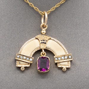 Può includere: Una collana con un ciondolo in oro decorato con una pietra preziosa viola e perle. Il ciondolo ha la forma di un arco con un motivo decorativo.