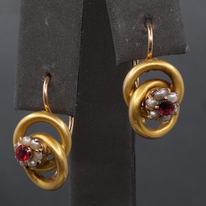 May include: A pair of gold-toned drop earrings featuring a circular hoop design. Each earring centers a cluster of small white seed pearls surrounding a single round, deep red gemstone. The earrings are suspended from curved gold-toned ear wires.
