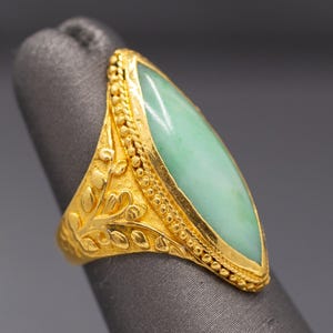 May include: A gold ring featuring a light green, elongated gemstone. The ring has intricate gold detailing, including floral and beaded patterns. The gemstone is set in a gold bezel, creating a striking contrast.