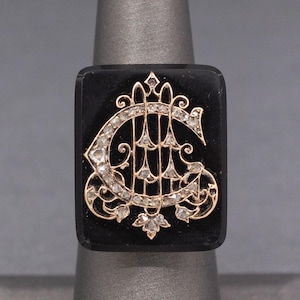 以下が含まれることがあります： ダイヤモンドが散りばめられたモノグラムが施された、金色のセッティングが施された黒オニキスのリング。モノグラムは装飾的な花のデザインで囲まれています。