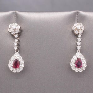 May include: A pair of elegant drop earrings. Each earring features a flower-shaped top, a series of small round stones, and a pear-shaped pendant with a deep red stone surrounded by smaller white stones. The earrings are set against a gray background.