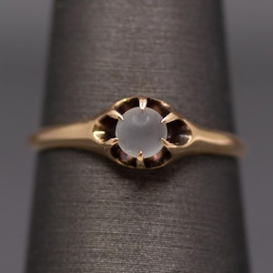 Könnte beinhalten: Ein goldener Ring mit einem zentralen, runden, durchscheinenden, mattweißen Stein, der von vier spitzen Krappen gehalten wird. Die Metallschiene ist glatt und verjüngt sich nach hinten, wobei die Fassung einen markanten, offenen, klauenartigen Rahmen bildet.