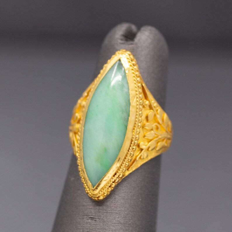 May include: A gold ring featuring a marquise-cut, light green jade stone. The ring has an ornate, filigree design with floral accents on the sides. The setting and band are a bright, polished gold color.