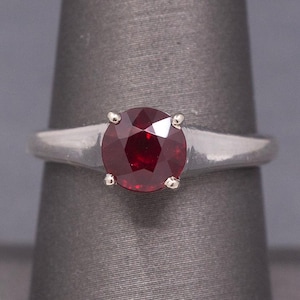 May include: A silver ring with a large, round, deep red gemstone. The gemstone is secured by four small prongs. The ring's band is smooth and narrows towards the back.