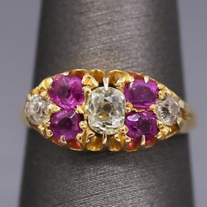 May include: A gold ring featuring a central cushion-cut white diamond flanked by four round-cut deep pink rubies and two smaller round-cut white diamonds on the outer edges, all set in a yellow gold band with a textured, scalloped metal setting.