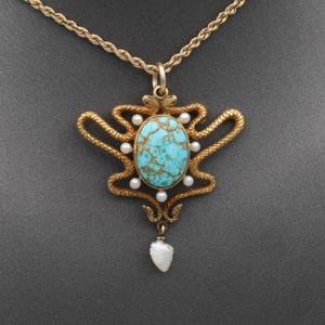 Op de afbeelding: Een goudkleurige ketting met een centrale ovale turkoois cabochon met bruine matrix, omringd door een gestructureerd slangenmotief. De hanger is versierd met kleine witte parels en een druppelvormige parel aan de onderzijde.