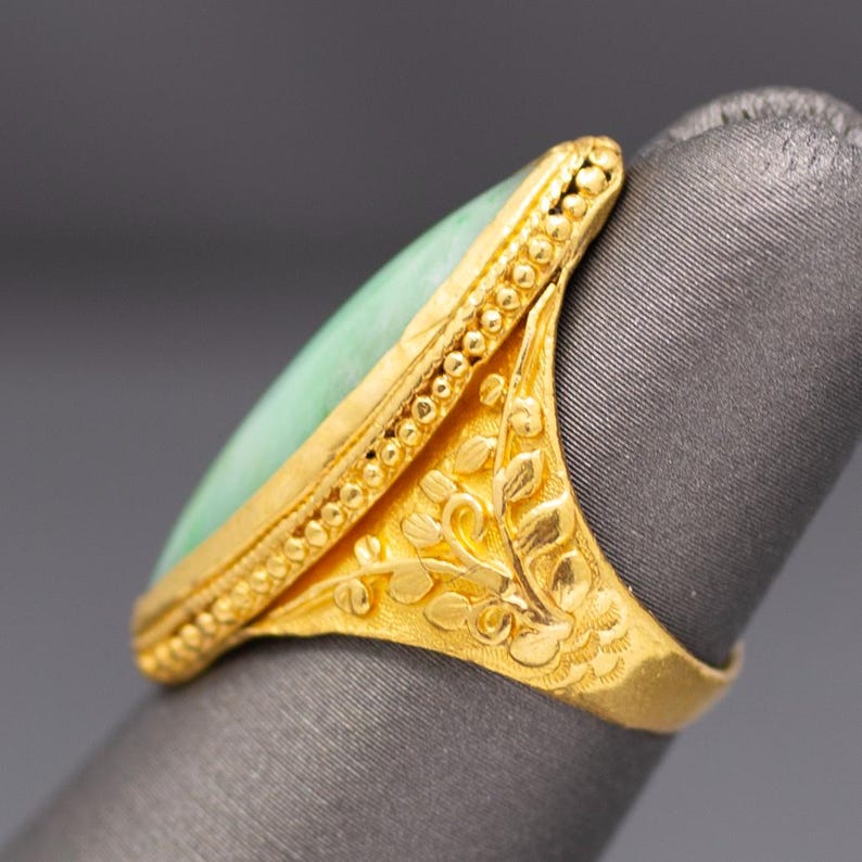 May include: A gold ring with a light green, elongated stone. The ring features intricate floral and dot patterns on the band and around the stone. The ring is set on a gray surface.