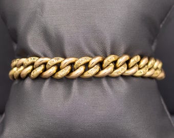 May include: A gold-colored chain bracelet with a series of interlocking oval links. Each link features an engraved design. The bracelet is displayed on a dark gray surface.