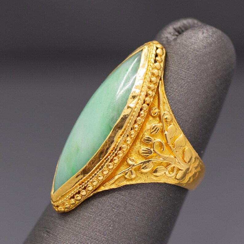 May include: A gold ring featuring a large, oval-shaped, pale green jade stone. The ring has intricate gold detailing, including a beaded border around the jade and floral patterns on the band. The ring is displayed on a gray surface.