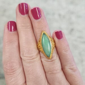 May include: A gold ring with an elongated, light green stone. The ring has intricate filigree detailing around the stone and on the band. The background shows a hand with burgundy nail polish.