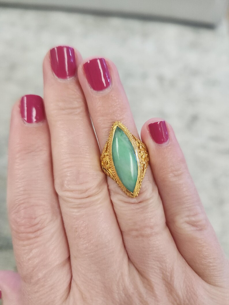 May include: A gold ring with an elongated, light green stone. The ring has intricate filigree detailing around the stone and on the band. The background shows a hand with burgundy nail polish.