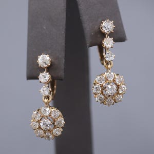 May include: A pair of gold drop earrings featuring a vertical row of three round faceted clear stones leading to a circular cluster of ten smaller round faceted clear stones surrounding a central round faceted clear stone in each earring.
