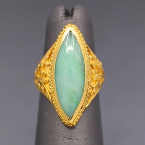 May include: A gold ring with a light green, oval-shaped stone. The ring has intricate floral designs on the sides and a beaded border around the stone. The ring is displayed on a gray finger.