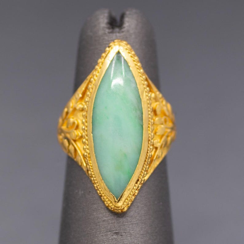 May include: A gold ring with a light green, oval-shaped stone. The ring has intricate floral designs on the sides and a beaded border around the stone. The ring is displayed on a gray finger.