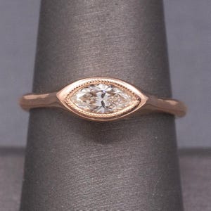 May include: Rose gold ring featuring a marquise-cut diamond. The diamond is set in a bezel with a delicate beaded edge. The band is smooth and rounded. The ring is displayed on a gray background.