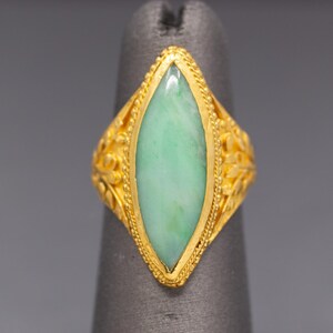 May include: A gold ring featuring a light green jade stone in a marquise shape. The ring has a detailed floral design on the sides and a beaded border around the stone. The ring is displayed on a gray background.