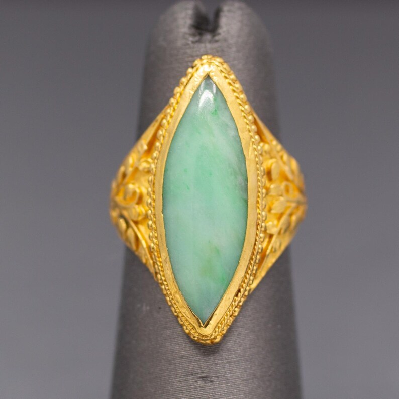 May include: A gold ring featuring a light green jade stone in a marquise shape. The ring has a detailed floral design on the sides and a beaded border around the stone. The ring is displayed on a gray background.
