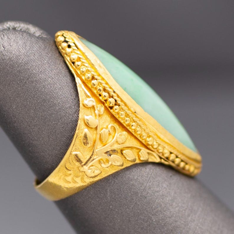 May include: A gold ring with a light green, oval-shaped stone. The ring has intricate floral and beaded detailing. The band and setting are gold. The ring is shown on a gray surface.