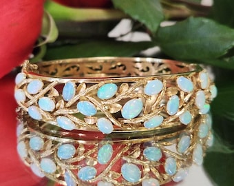 May include: A gold-toned hinged bangle bracelet featuring an openwork design of intertwined branches and leaves. The bracelet is adorned with numerous oval-shaped opals in shades of blue and white. The bracelet is reflected in a mirrored surface.