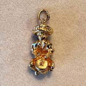 May include: A gold-toned metal charm depicting a figure wearing a wide-brimmed hat, seated while playing a drum. The charm features a circular jump ring at the top for attachment. The figure is rendered in a metallic gold finish with detailed textures.
