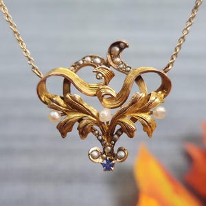 Può includere: Una collana con pendente dorato con un design ornato e a spirale. Il pendente presenta piccole perle e una gemma blu. La collana ha una catena delicata ed è probabilmente vintage o antica.