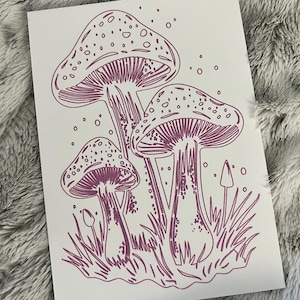 May include: A white print featuring a cluster of stylized purple mushrooms with dotted caps and detailed gills. The illustration includes grass and small dotted accents, creating a whimsical, nature-inspired design.