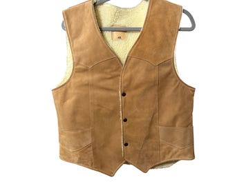 May include: A tan suede vest with a cream-colored shearling lining. The vest has a V-neck, two front pockets, and three buttons. It is hanging on a gray hanger against a white background.