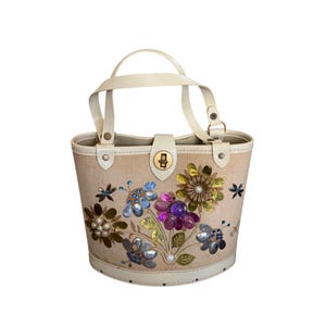 May include: A beige handbag with a cream-colored handle and trim. The bag is decorated with colorful floral embellishments, including blue, purple, and gold gemstones. It has a gold-colored clasp.