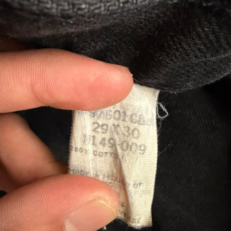 May include: Close-up of a tag from a pair of black denim jeans. The tag is off-white and features text including the size 29x30, and the material 100% cotton. The tag is partially obscured by a hand.