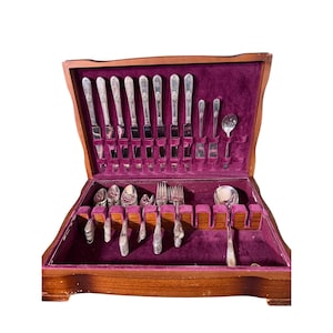 May include: A wooden flatware chest with a deep purple velvet interior containing a set of silver-toned metal utensils. The collection includes various knives, forks, and spoons arranged in designated slots within the open storage box.
