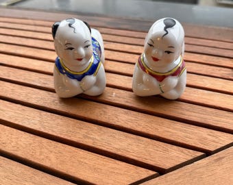May include: Two small, white porcelain figurines of seated figures with black hair and red lips. One wears a blue and yellow outfit, the other a red and yellow outfit. The figurines are on a wooden surface.
